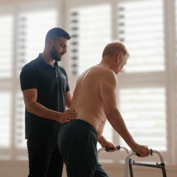 NDIS-registered physiotherapist providing hands-on mobility support and gait retraining to an older male participant using a walking frame during an in-home physiotherapy session in Melbourne.